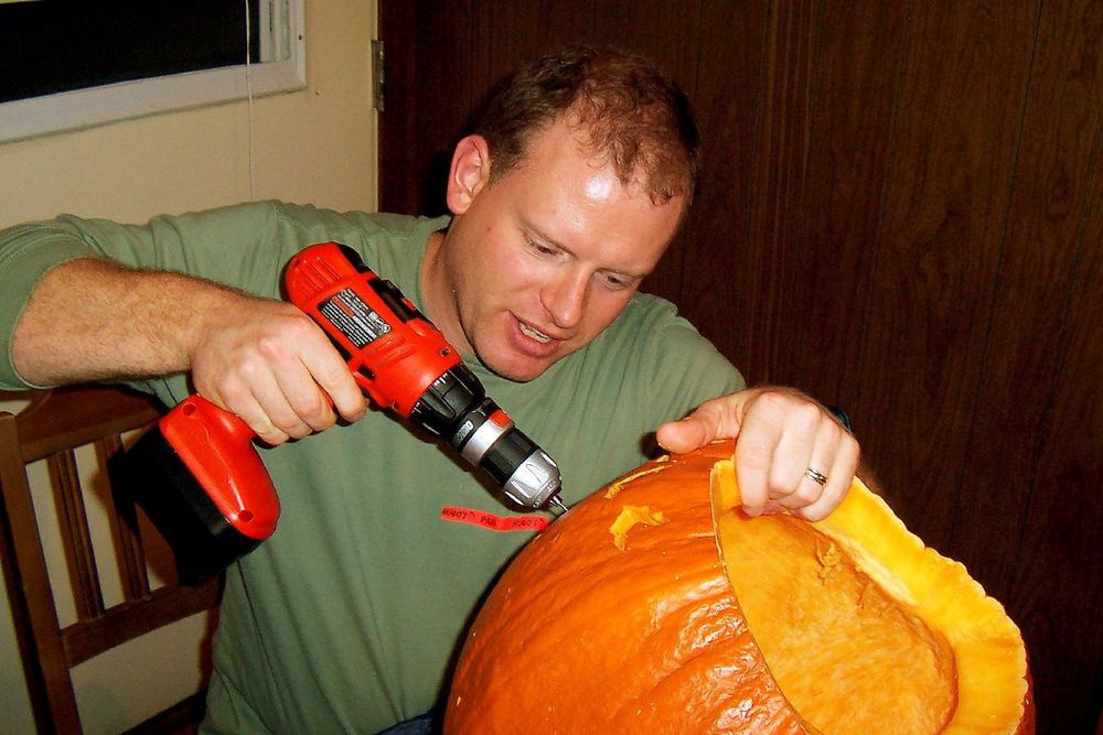 A man using a power drill to carve a pumpkin for Halloween