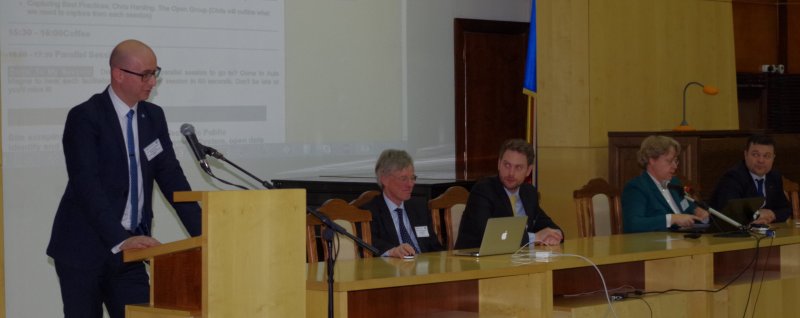 A man at a lectern addressing an audience, members of the panel are looking on