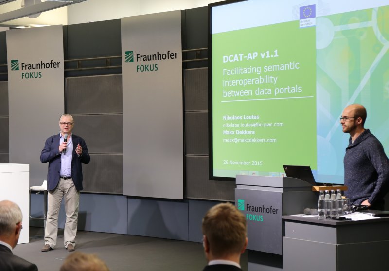 Two men on the podium at Fraunhofer FOKUS, Nikos Loutas stands in fromt of a screen on which his title slide can be seen (DCAT-AP 1.1) Facilitating Interoperability between data portals
