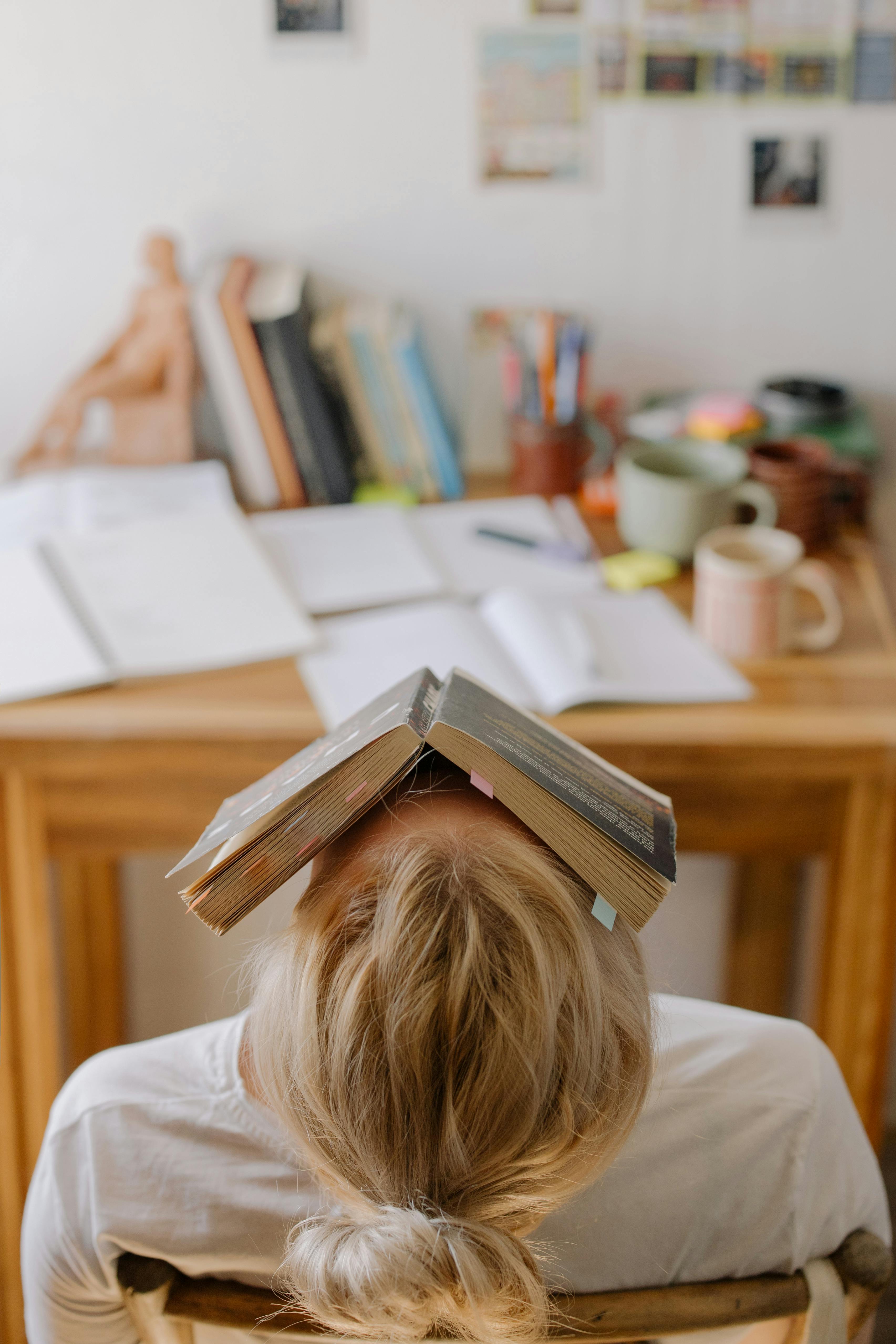 A woman sitting at a cluttered desk, she is leaning back in her chair towards the camera with a book over her face. Photo by cottonbro studio.