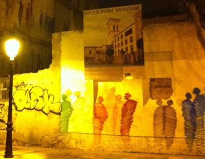 Shadow like figures and a graffiti tag drawn on the walls of a partially demolished building, illuminated by the light from a street lamp.