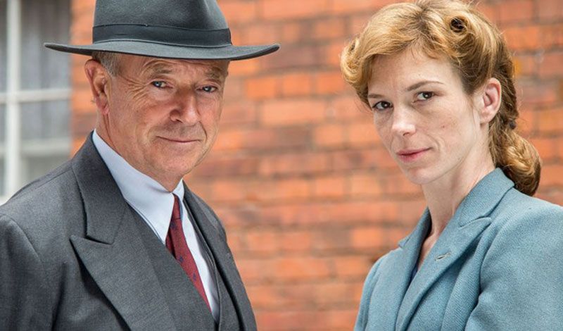 A man in a suit and fedora and a woman with coiffed hair look sternly into the camera.