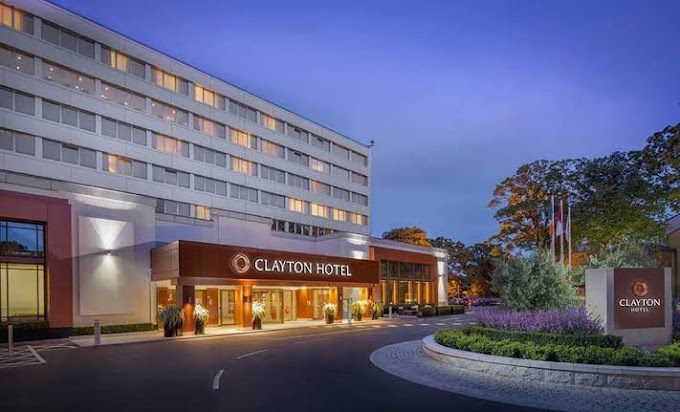 Street view at dusk of the facade of the Clayton Hotel and its driveway. The building has six stories. Some windows are lit.