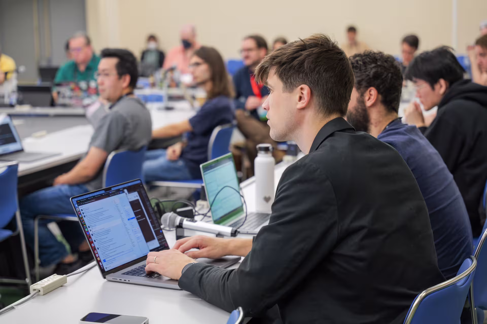 Participants at a conference meeting seated at desks arranged in a U shape. Some are typing on laptops, and some have their heads turned towards a speaker off camera.